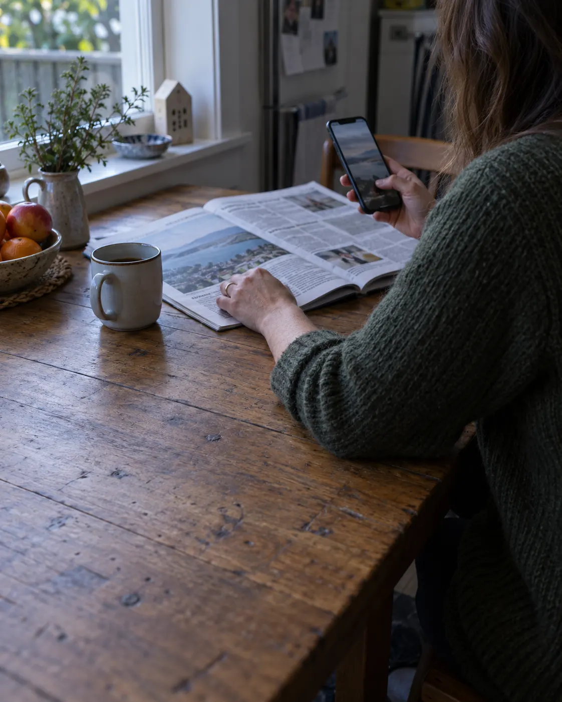 A person at a kitchen table reading the newspaper and looking at their phone